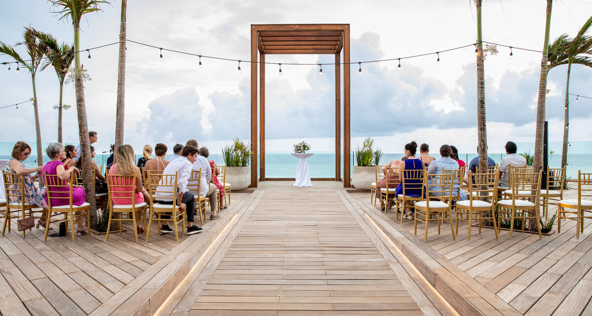 A beachfront wedding ceremony setup with gold chairs and an elevated wooden arch framing the view of the ocean. Guests are seated, and string lights are hung above the aisle. Keywords: destination wedding planning, luxury destination weddings, oceanfront wedding.