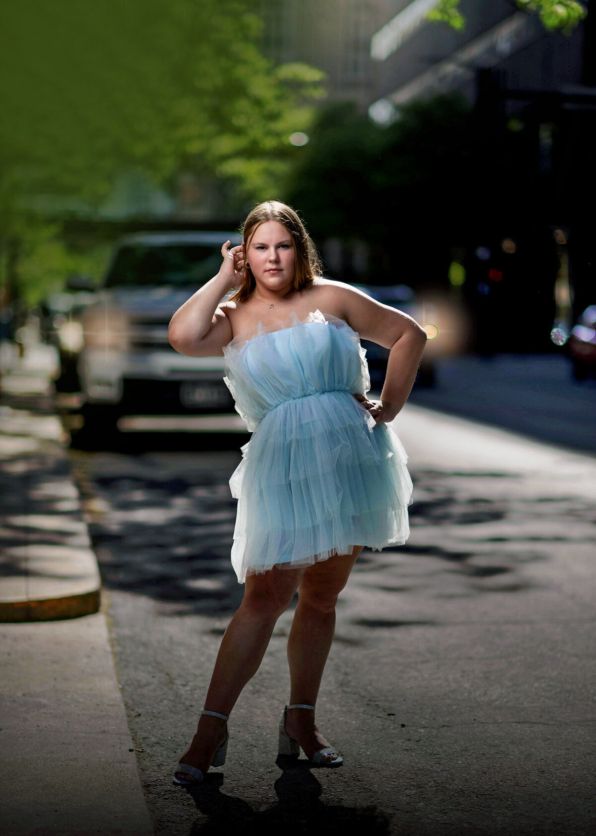 A high school senior stands in a downtown street with a hand in her hair and on her hip in a blue tulle dress