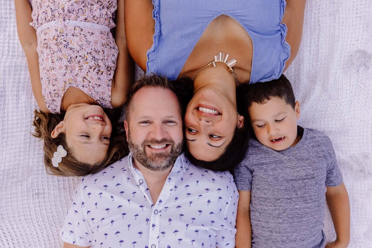 Family lying on a blanket outdoors, heads together and smiling joyfully at the camera in a playful pose.