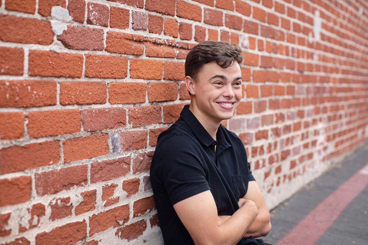 A person in a black shirt leans against a red brick wall with arms crossed, smiling and looking to the side. The wall extends into the distance, and the ground is partially visible.