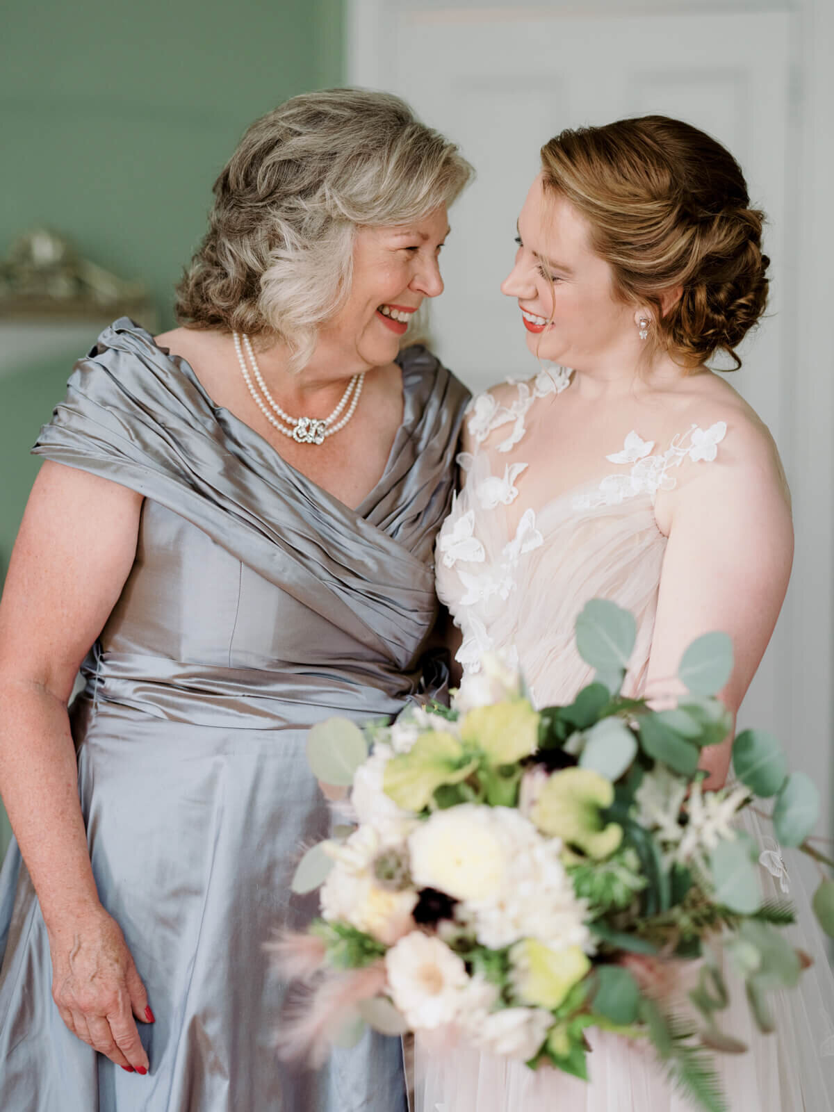 The bride, holding her flower bouquet, is standing side by side with an old lady while happily looking at each other.