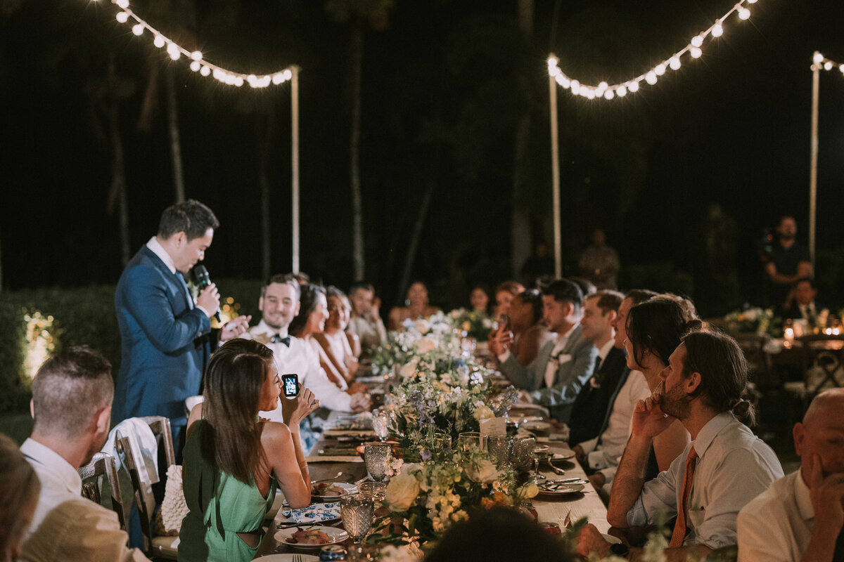 Ambient light of wedding reception guest standing up at table to give a speech while everyone watches