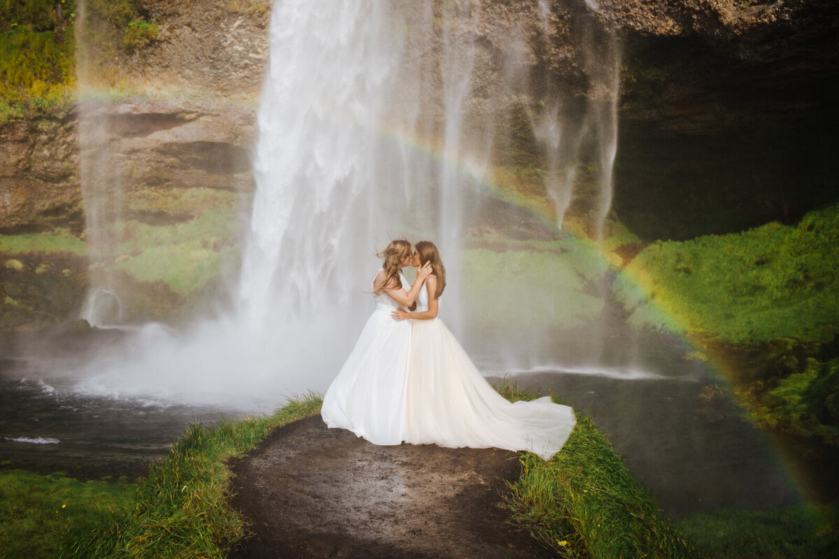 two brides kiss under waterfall rainbow in wedding gowns