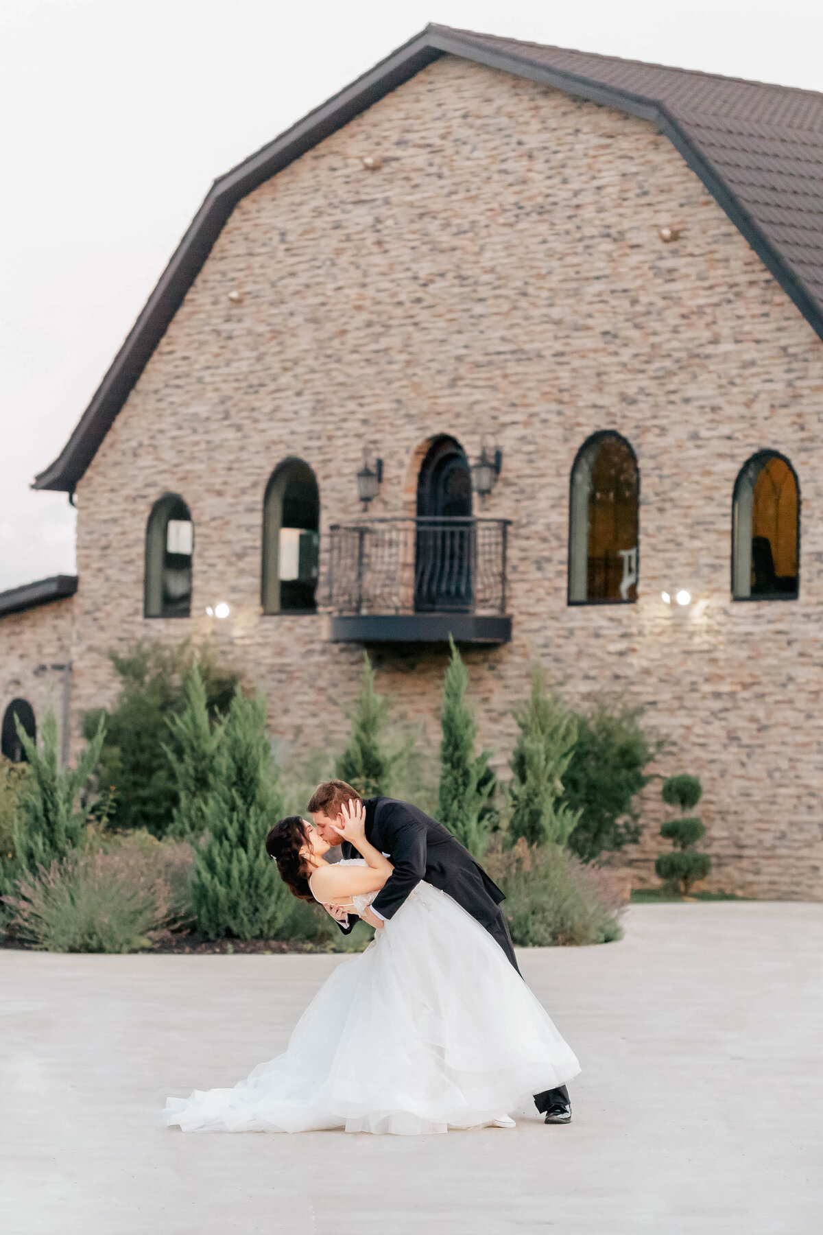 Bride and groom sharing a dip kiss outside a rustic stone chapel. The playful and romantic moment is captured with the chapel’s architecture as a charming backdrop, adding to the timeless feel of the photo.