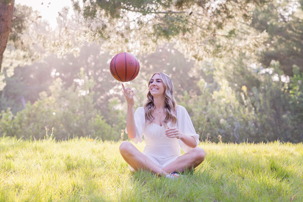 A person with long hair sits cross-legged on a grassy field, happily spinning a basketball on their finger. Sunlight filters through the trees in the background.