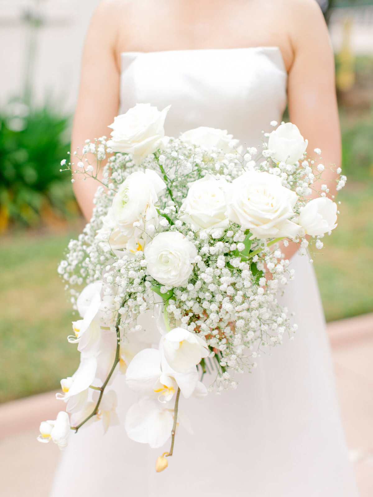 Bride in a strapless white gown holding a cascading bouquet of white roses, baby's breath, and orchids, set against a green garden backdrop. Elegant and serene.