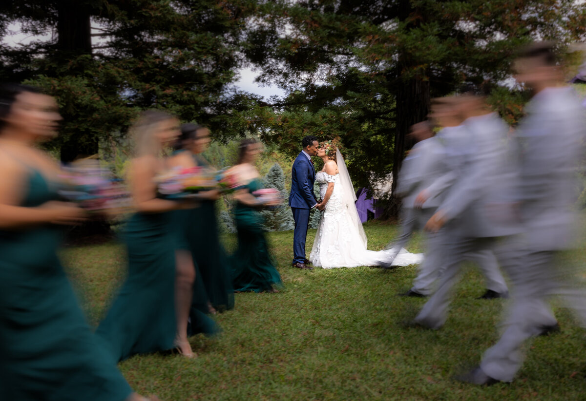Bride and groom stand in middle while bridesmaids and groomsmen walk in front of them, blurred in camera. Creative wedding photo captured by sacramento wedding photographer philippe studio pro.