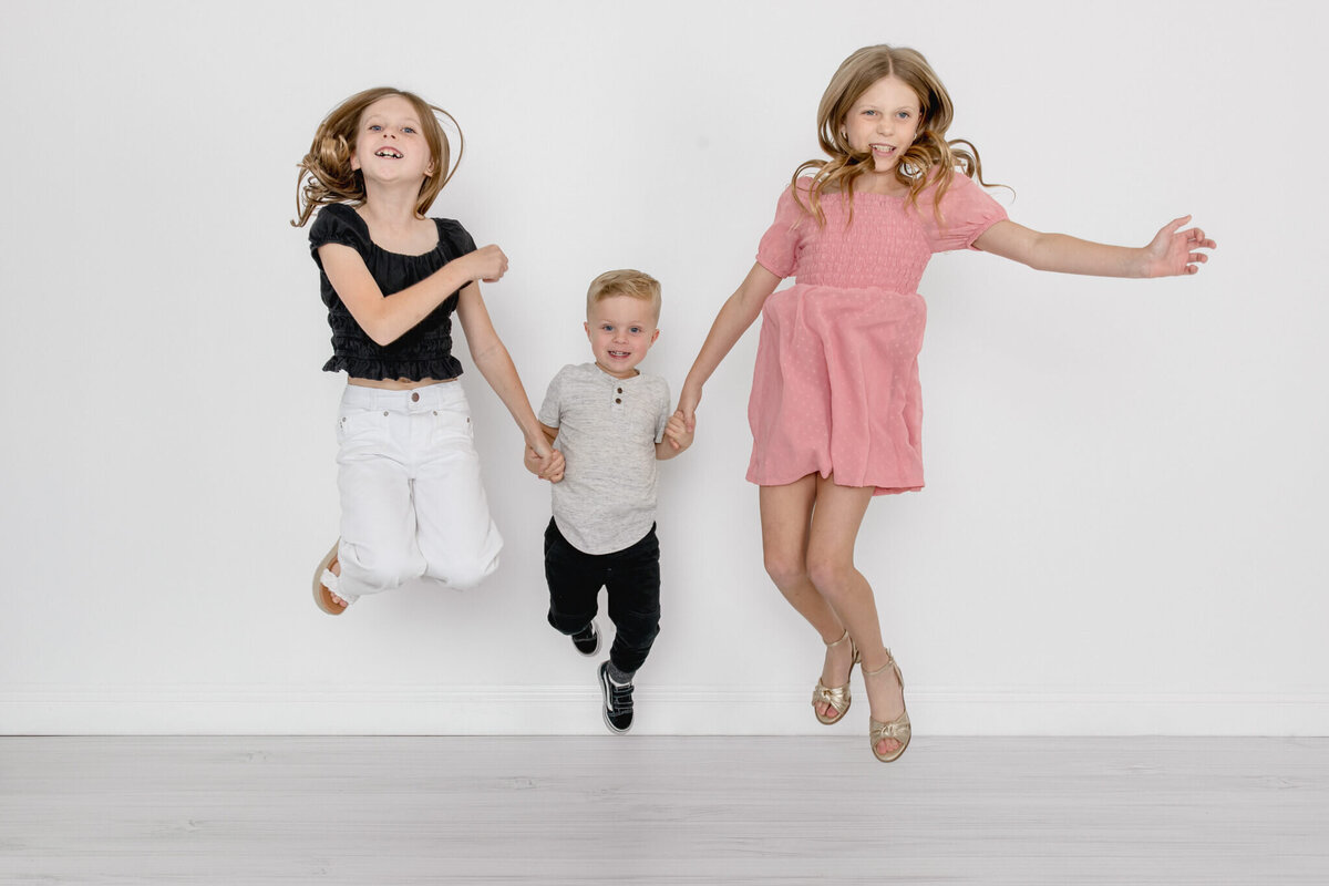 Two young girls with long blonde hair wearing white, pink, and black, holding their little brother's hands on either side of him, jumping in the air and laughing, looking at the camera.