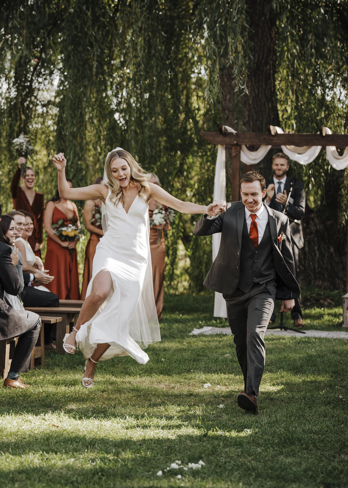 A joyful bride jumping kicking for joy and groom holding hands and running, with smiles on their faces, as guests cheer them on in an outdoor wedding setting taken by jen Jarmuzek photography a Minneapolis wedding photographer