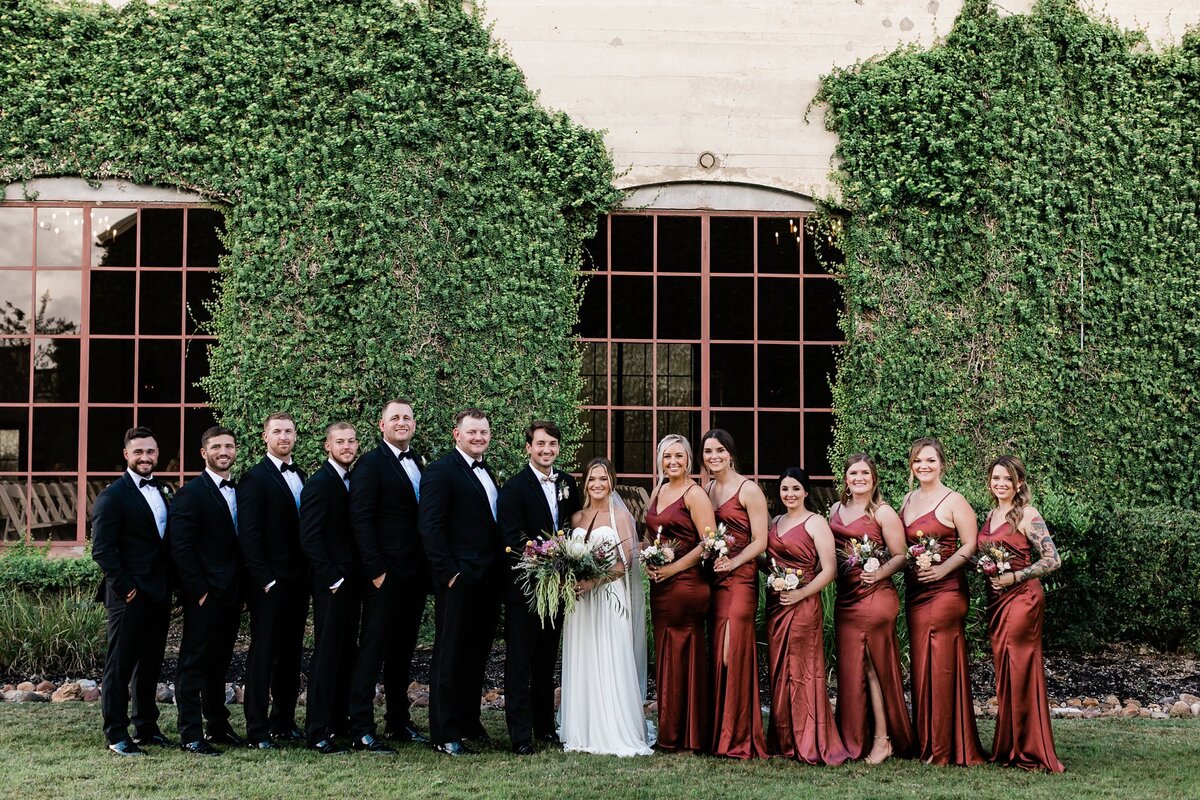 Wedding party portraits smiling at the camera in front of the chapel at Olde Dobbin Station. The chapel has green vines growing all along the wall as the backdrop.