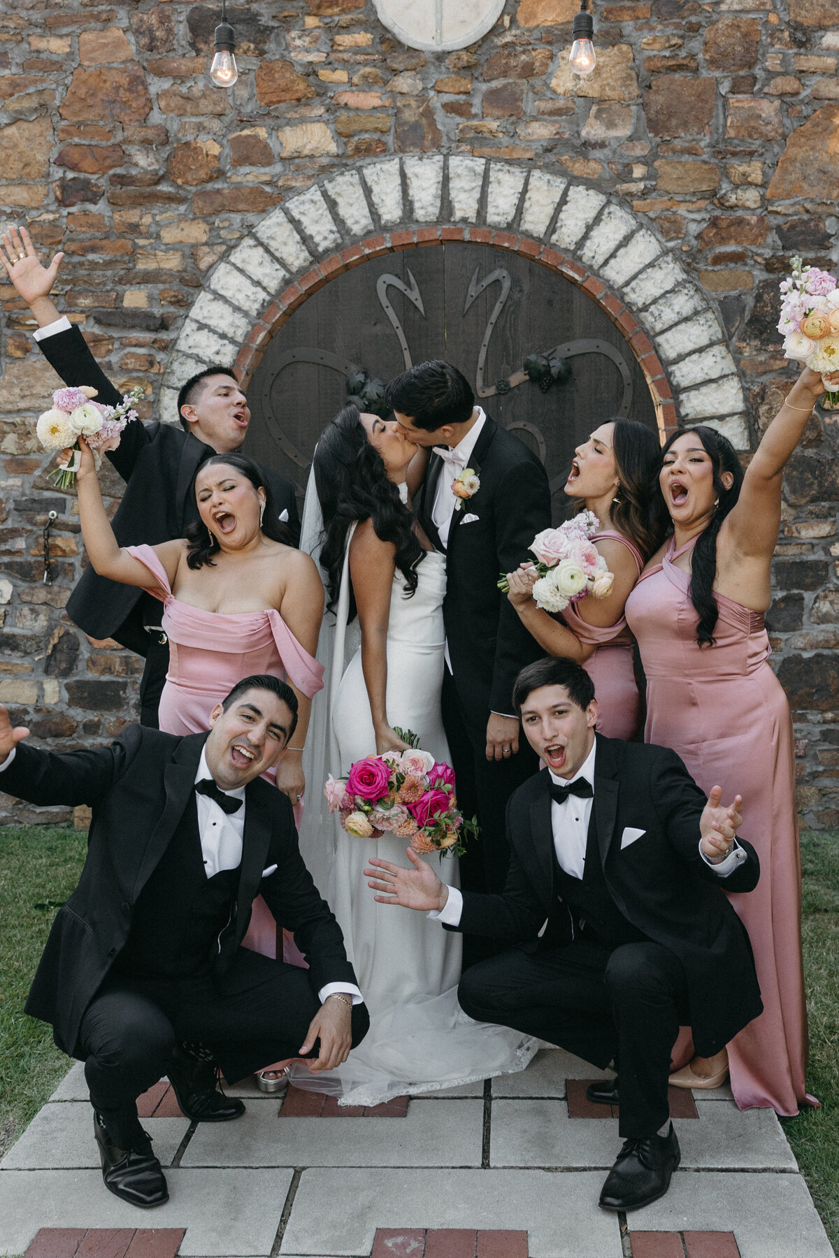 Playful wedding party celebration with bridesmaids in blush gowns and groomsmen in black tuxedos gathered around the newly married couple, highlighting vibrant garden-style bouquets with roses, dahlias, and ranunculus.
