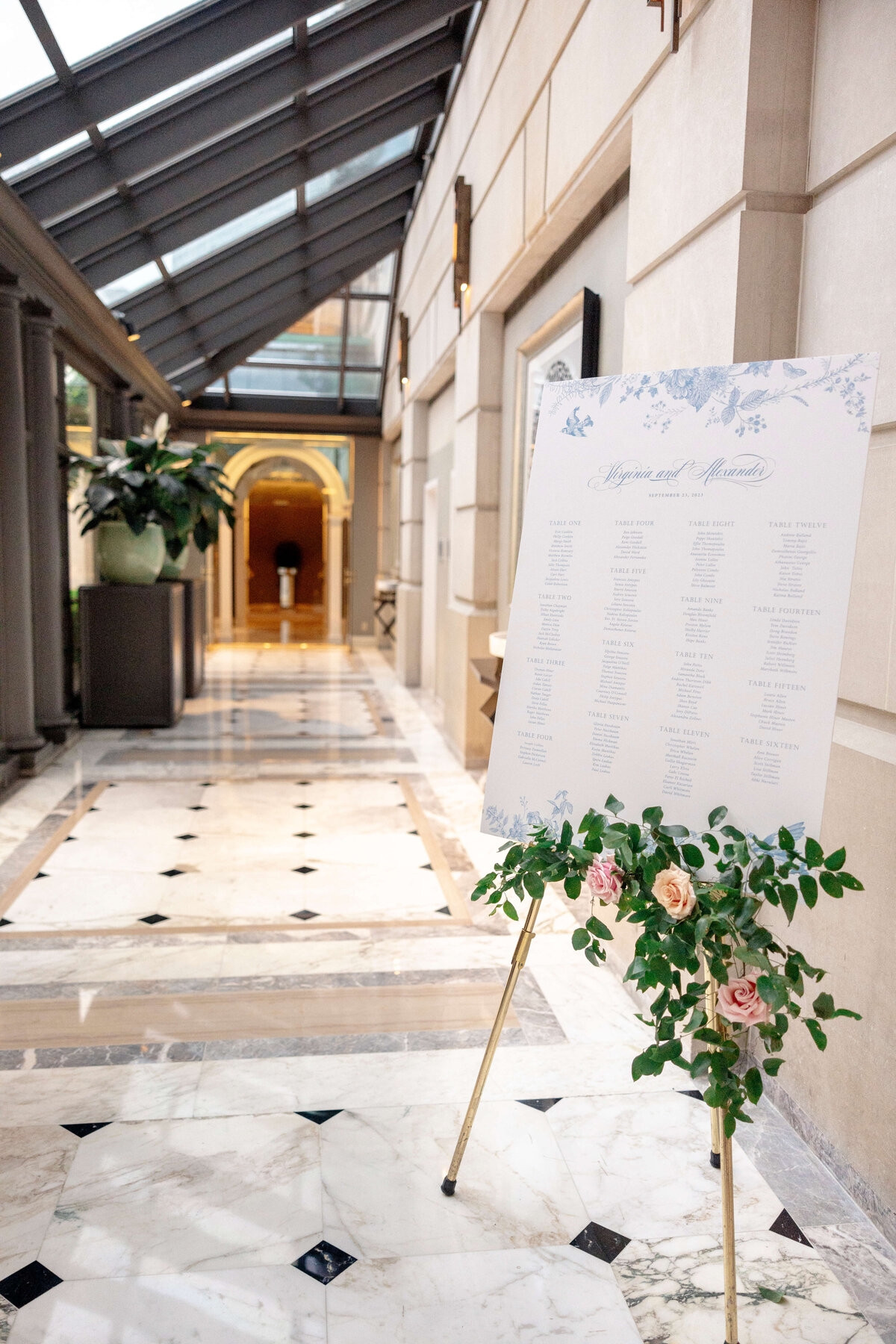 A wedding seating chart on an easel stands in a luxurious marble hallway. The chart is decorated with floral designs and greenery. The hallway has a glass ceiling, ornate columns, and a large potted plant near the entrance.