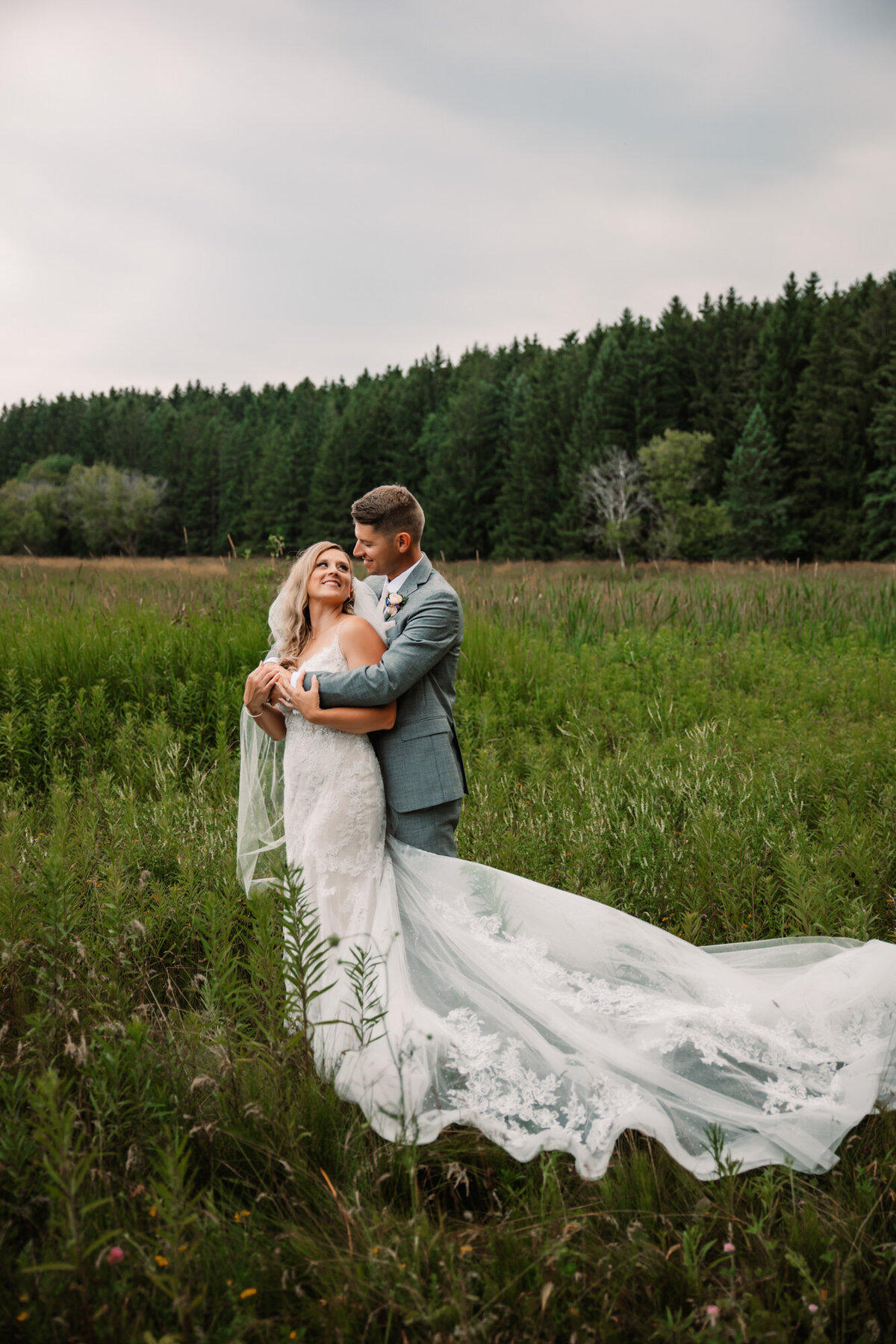 groom wraps his arm around bride from behind in a field of green while brides dress flows in the grass