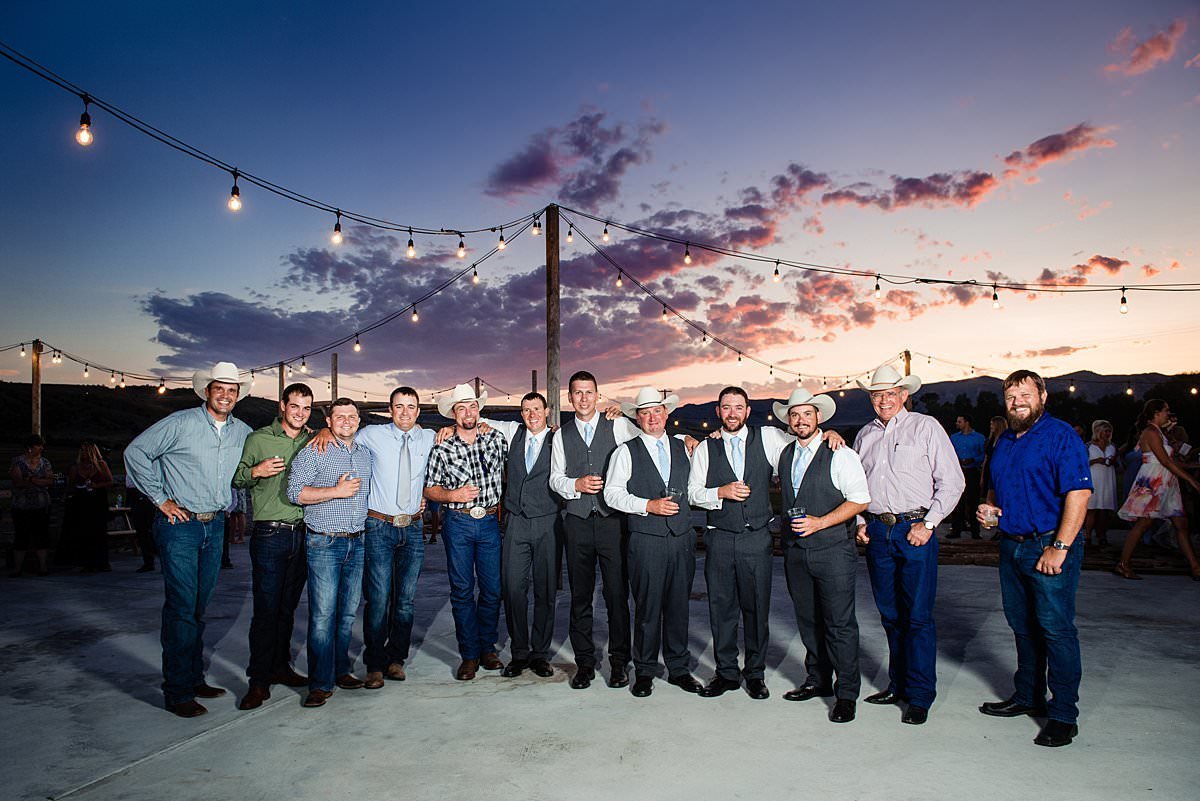 Group photo of Alpha Gamma Rho fraternity brothers together at their brothers wedding reception standing beneath a bright purple and orange Montana sunset