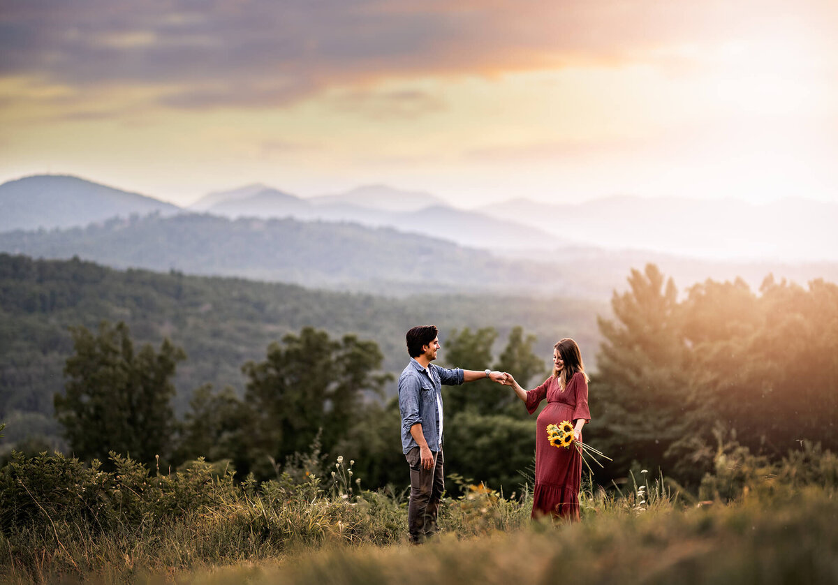 An expecting couple holds hands and dances in the setting sun with mountains in the distnace