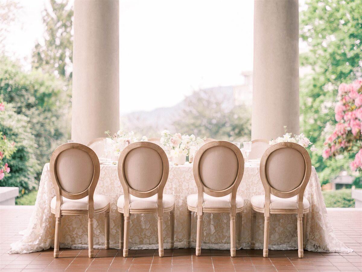 An elegantly set table outdoors with a lace tablecloth, six upholstered chairs, and floral arrangements, curated by Destination Wedding Planner Melissa Dawn Event Designs, positioned under a colonnade with lush greenery in the background.