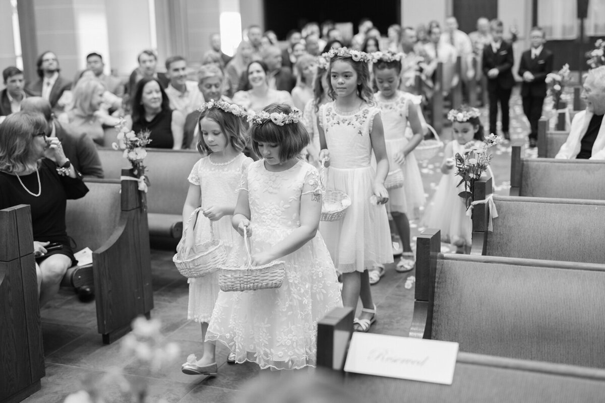 Black and white photo of eight flower girls walking down the aisle during a wedding ceremony.