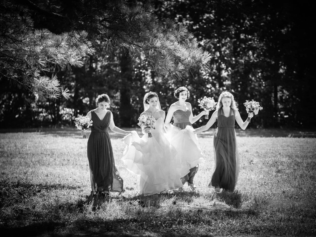 A black and white image captures the joyous motion of bridesmaids running through a field, surrounding the bride in her flowing gown.
