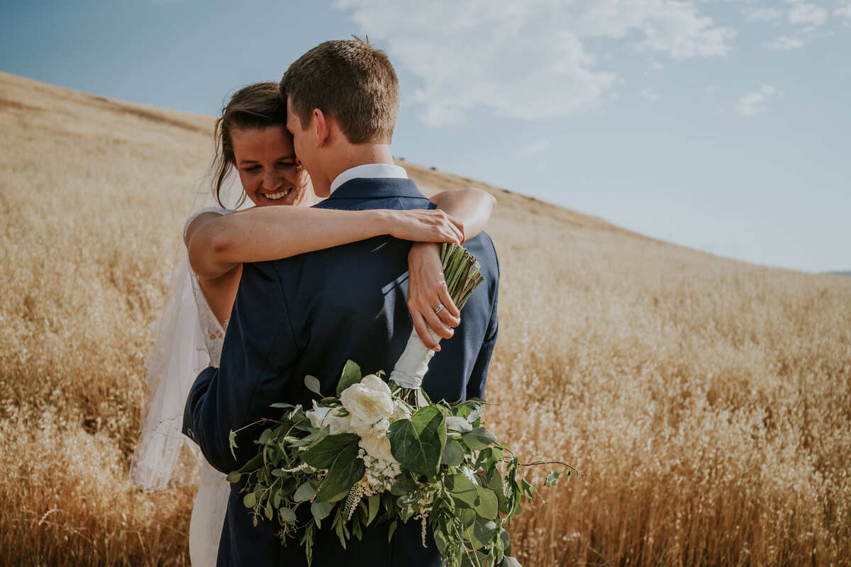 Bride hugs groom and holds bouquet behind his back while standing in golden field.