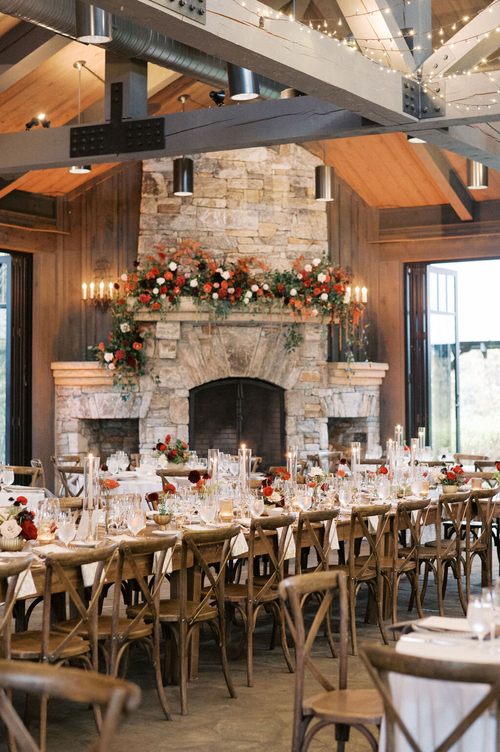 Long reception table with red flora bud vases and taper candles with stone fireplace decorated with red floral arrangements.