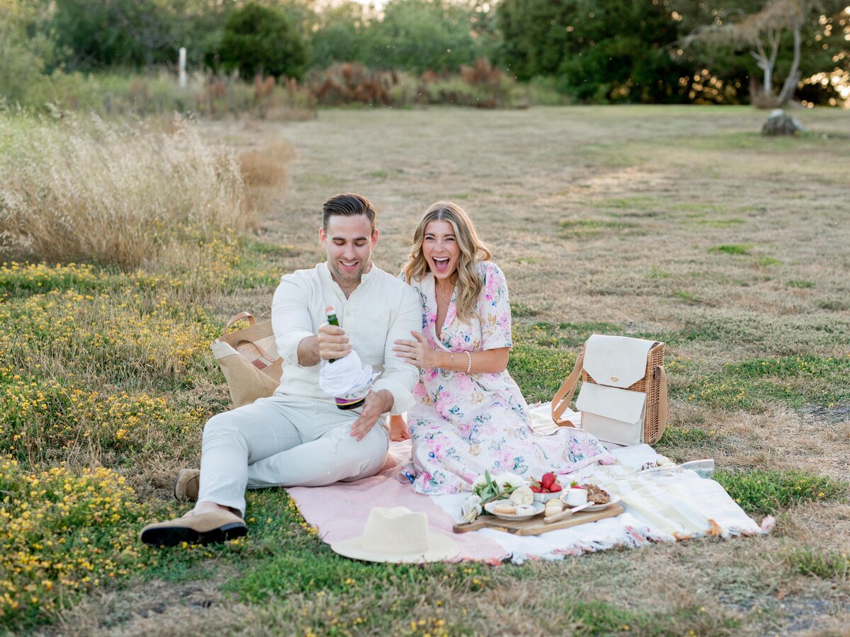 Champagne pop and pure joy—celebrating love with laughter during a romantic outdoor engagement picnic.