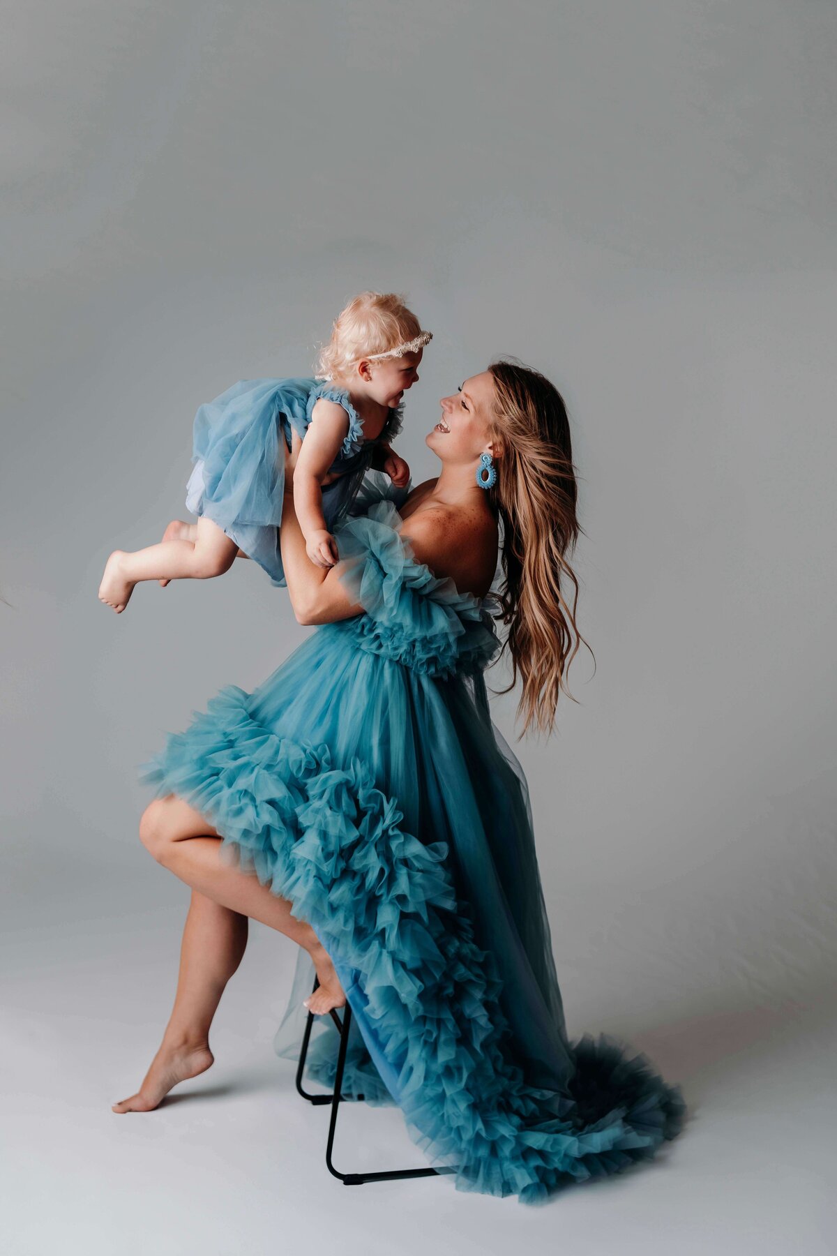 There's a young woman sitting on a stool, wearing a gorgeous blue tulle gown as she lifts her toddler daughter into the air for a playful, yet formal motherhood portrait by Mobile, AL Motherhood Photographer. The child is cheerful, wearing a matching blue tulle gown and a white floral headband.