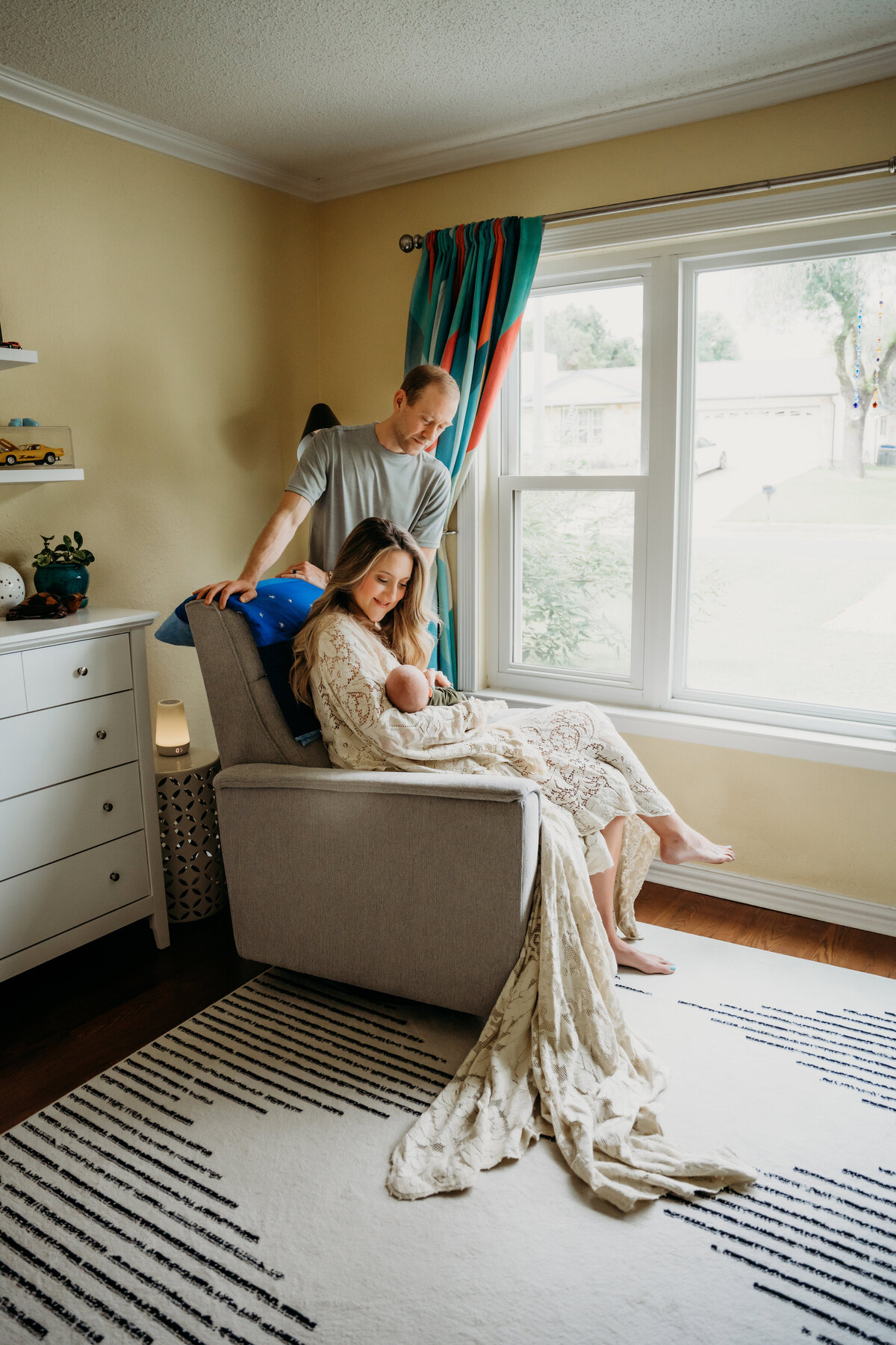 Newborn Photographer, a mother sits in a chair in the corner of her room with baby, dad leans over them in admiration