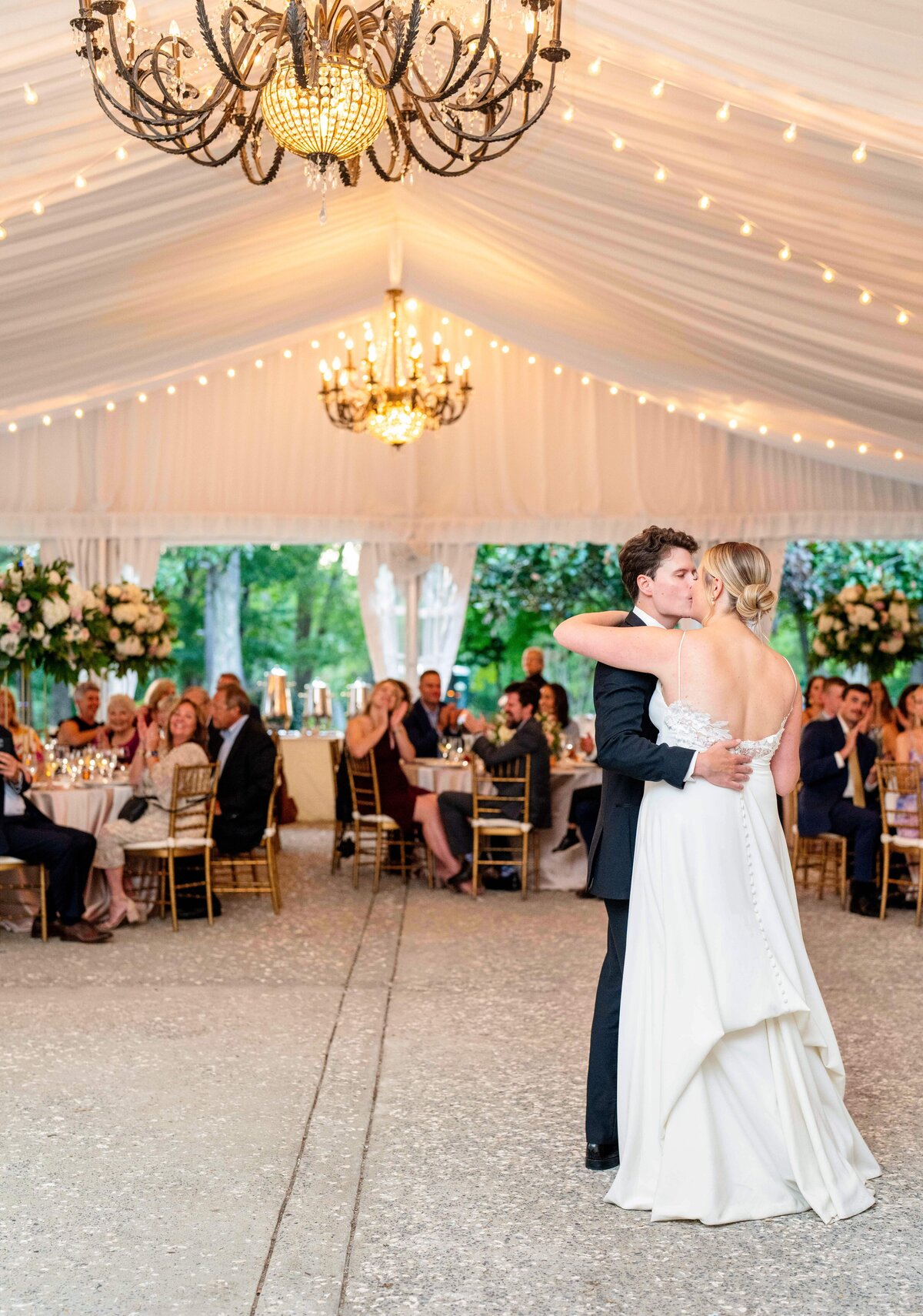 A couple shares a dance at a wedding reception under a white tent with chandeliers. Guests seated at tables with floral arrangements watch them. The bride wears a white gown, and the groom is in a dark suit. String lights illuminate the setting.