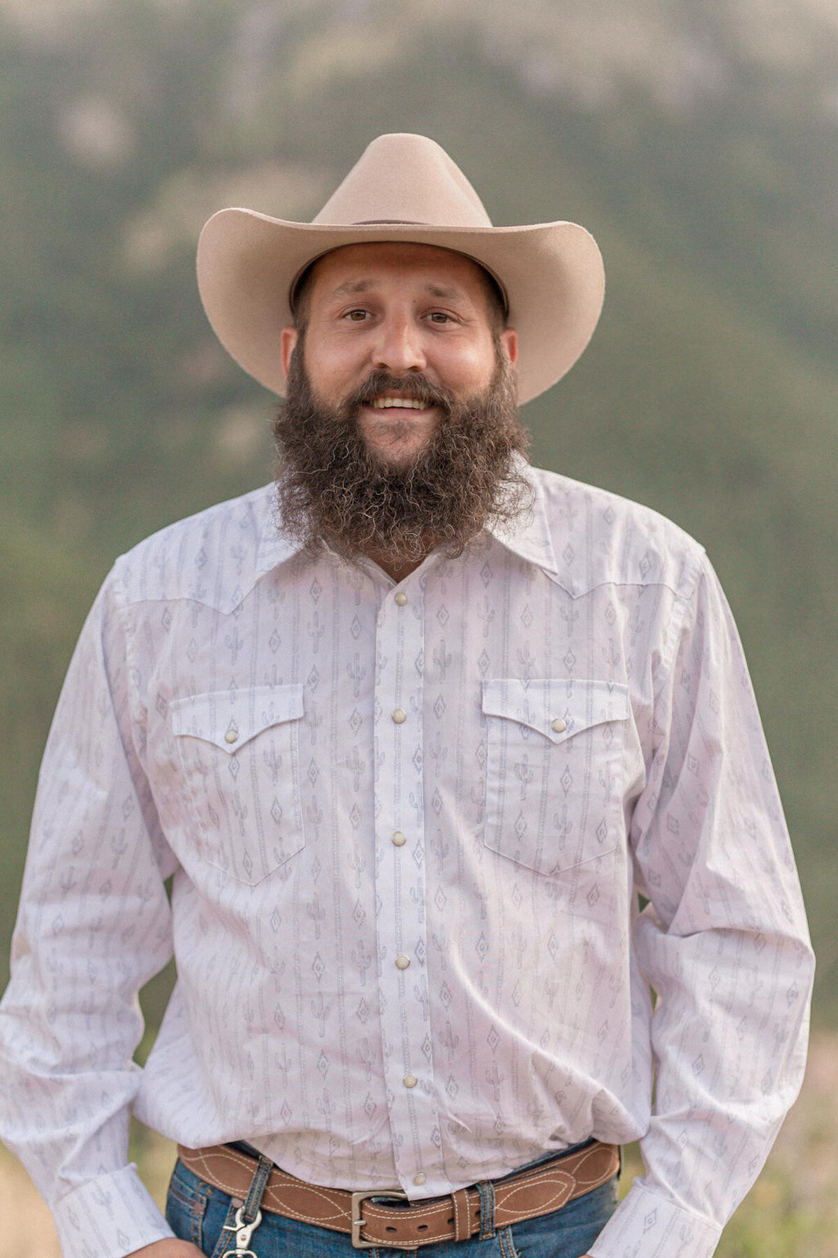 The groom stands, smiling at the camera, sporting a tan cowboy hat and bushy beard