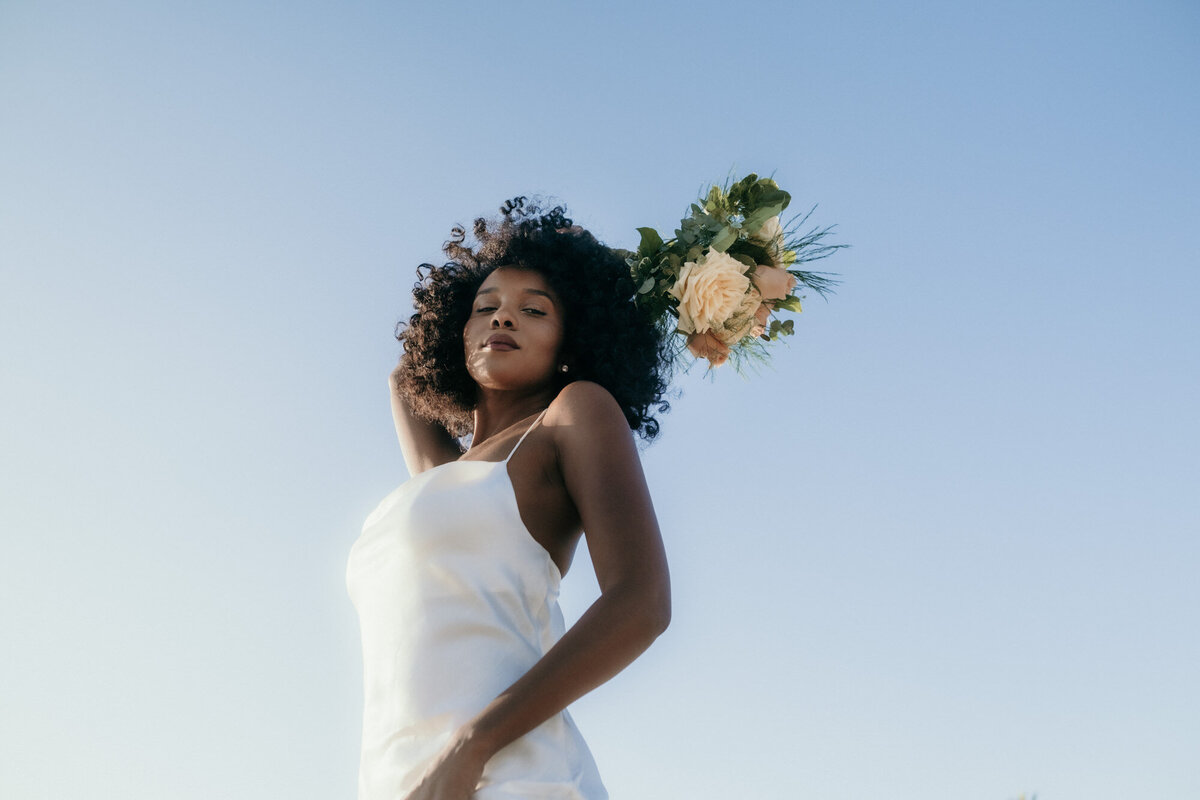 View from the bottom -the bride stands in the sunlight holding her bouquet behind her head, looking down at the camera.