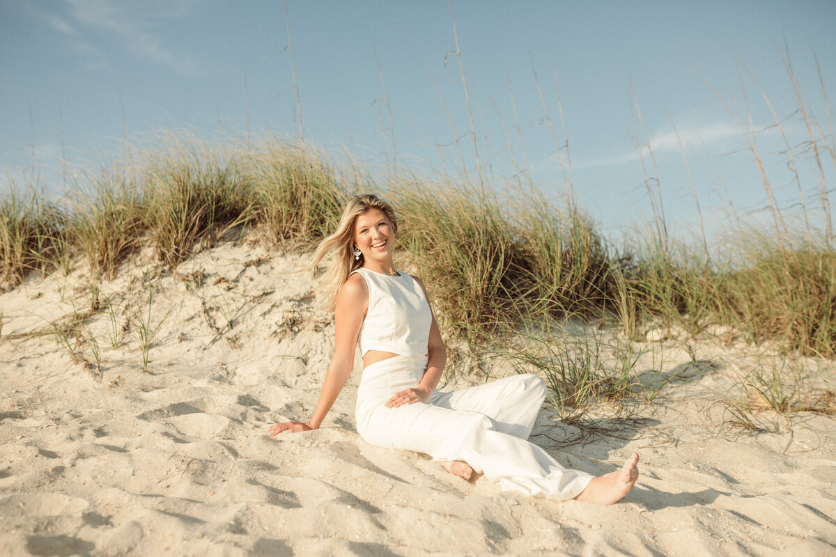 Senior portraits of high-school senior dressed in white linen jumpsuit poses in sand on Panama City Beach.  Portrait taken By Brittney Stanley of Be Seen Photos