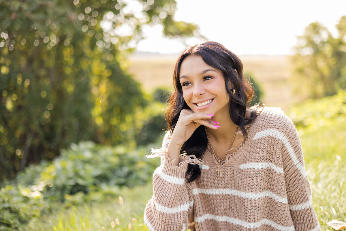 high school female senior, with long black hair, sitting in the grass, with trees behind her, looking away from the camera smiling, wearing a light tan and white stripped shirt