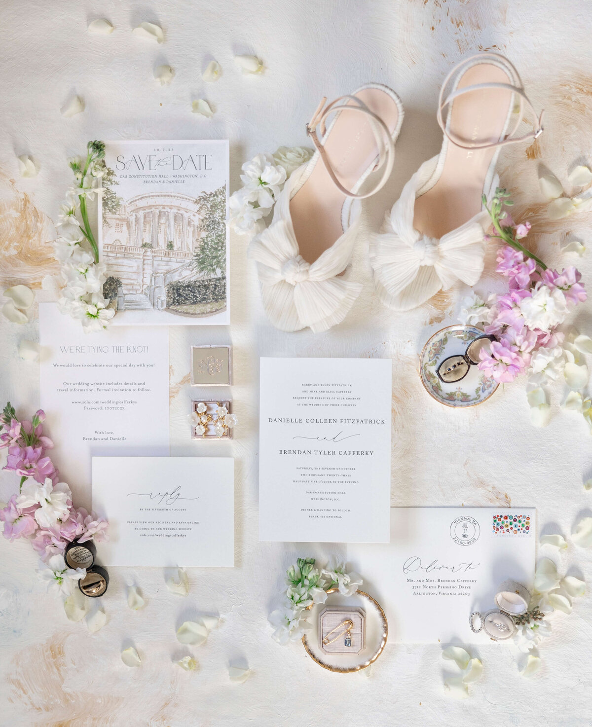 A flat lay of wedding items: white high-heel shoes with bows, a save-the-date card, invitations, jewelry, rings in a box, and floral accents. The scene is decorated with small white and purple flowers on a light surface.
