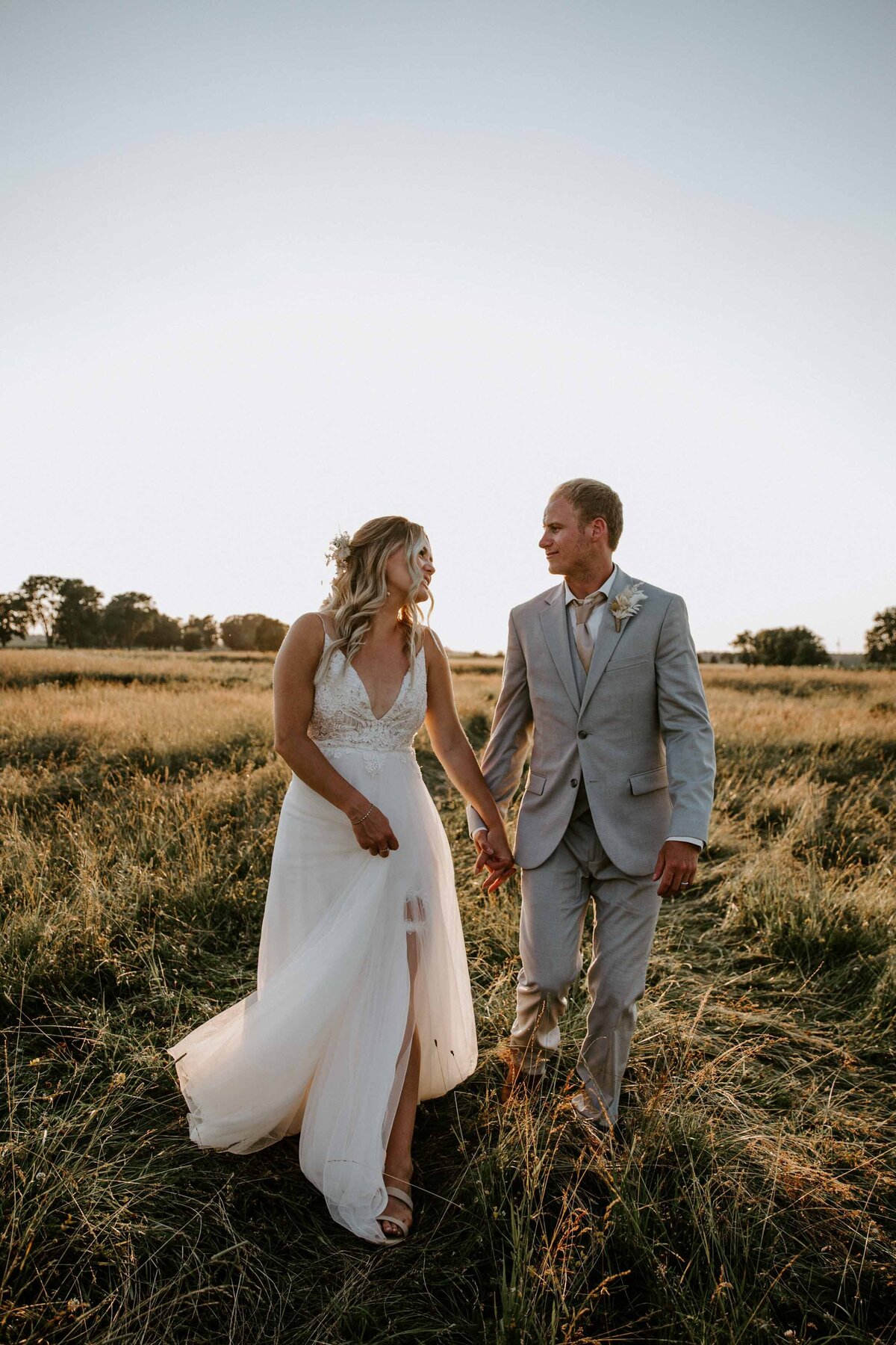 Bride and groom casually walk through an Exeter, Ontario field at their rustic barn wedding on family farm. Bride and groom are looking at each other with golden light wrapping around them.