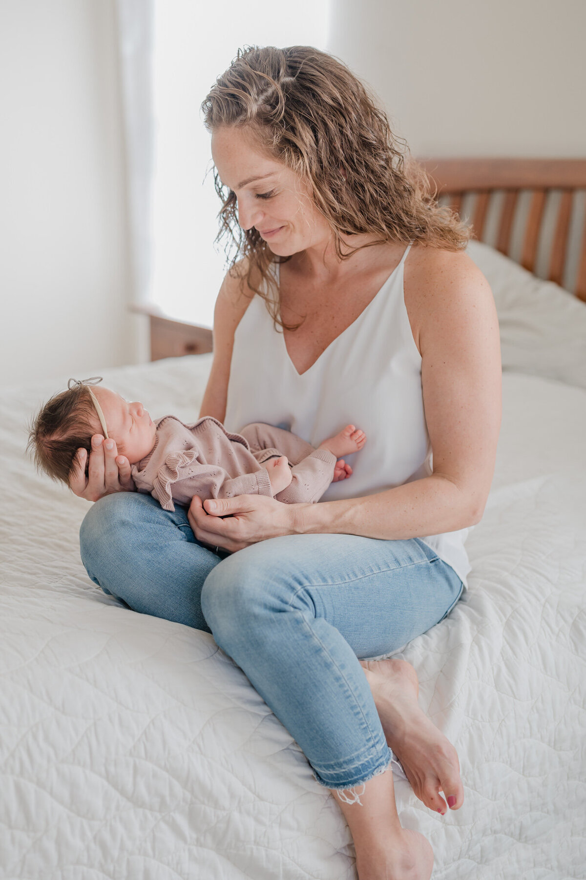 Mom sits on her bed and gazes down at her baby on her lap during lifestyle newborn pictures.