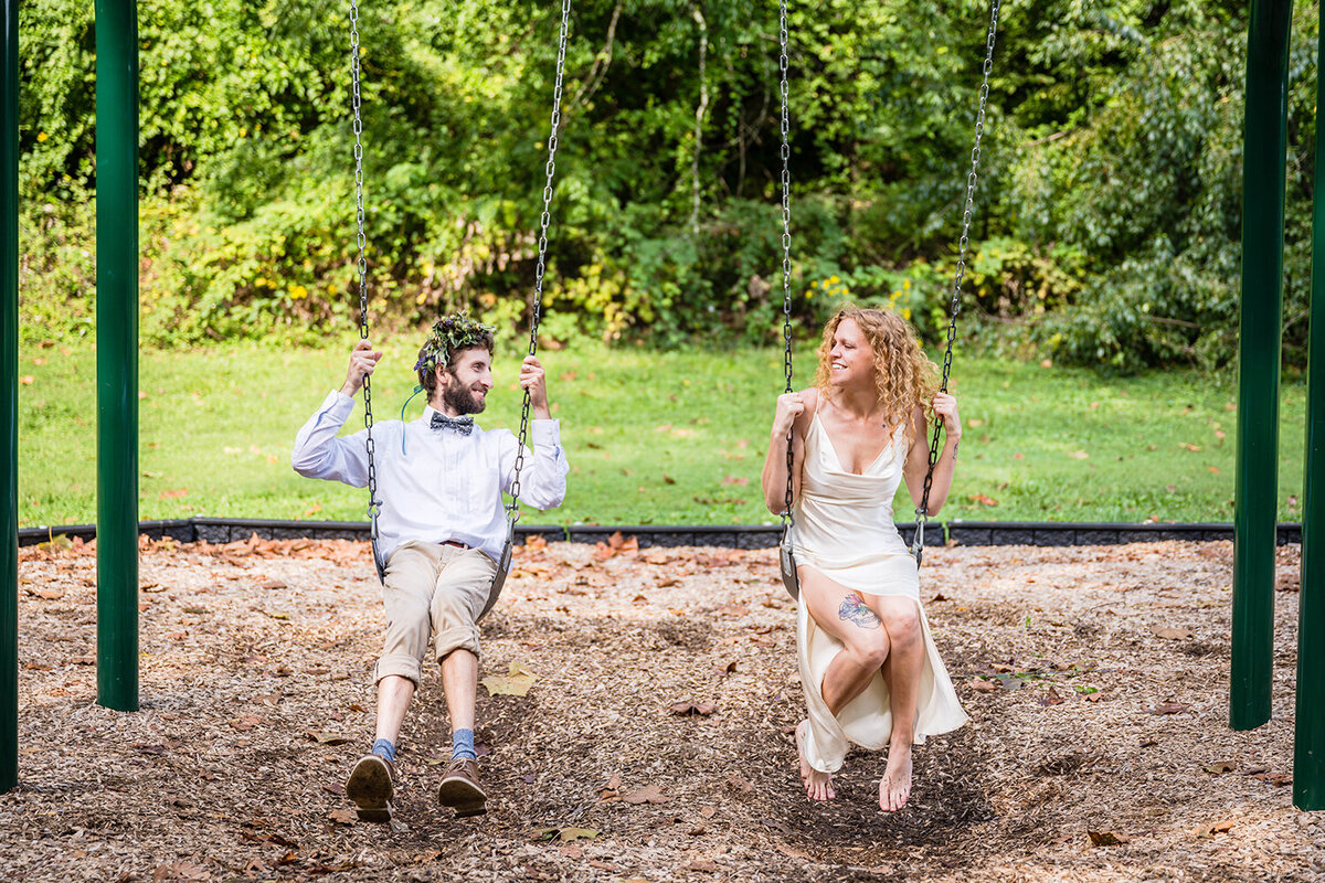 A bride and groom swing together on the swingset at Fishburn Park on their elopement day in Roanoke, Virginia and smile widely at one another.