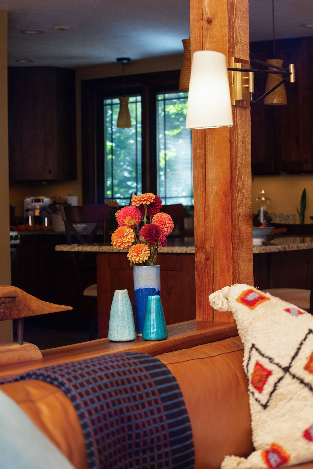 A detail shot highlighting the textures and colors of a cozy cabin living space. The smooth brown leather of the sofa contrasts with the woven throw and the textured pillow. Bright orange and red flowers in a white and blue vase add a pop of color, alongside small teal vases. Warm light emanates from a brass wall sconce.