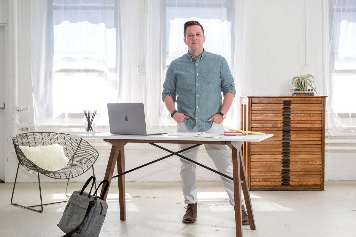 A male entrepreneur wearing business casual wear, standing behind his desk that has a laptop & papers on it, looking at the South Shore MA branding photographer