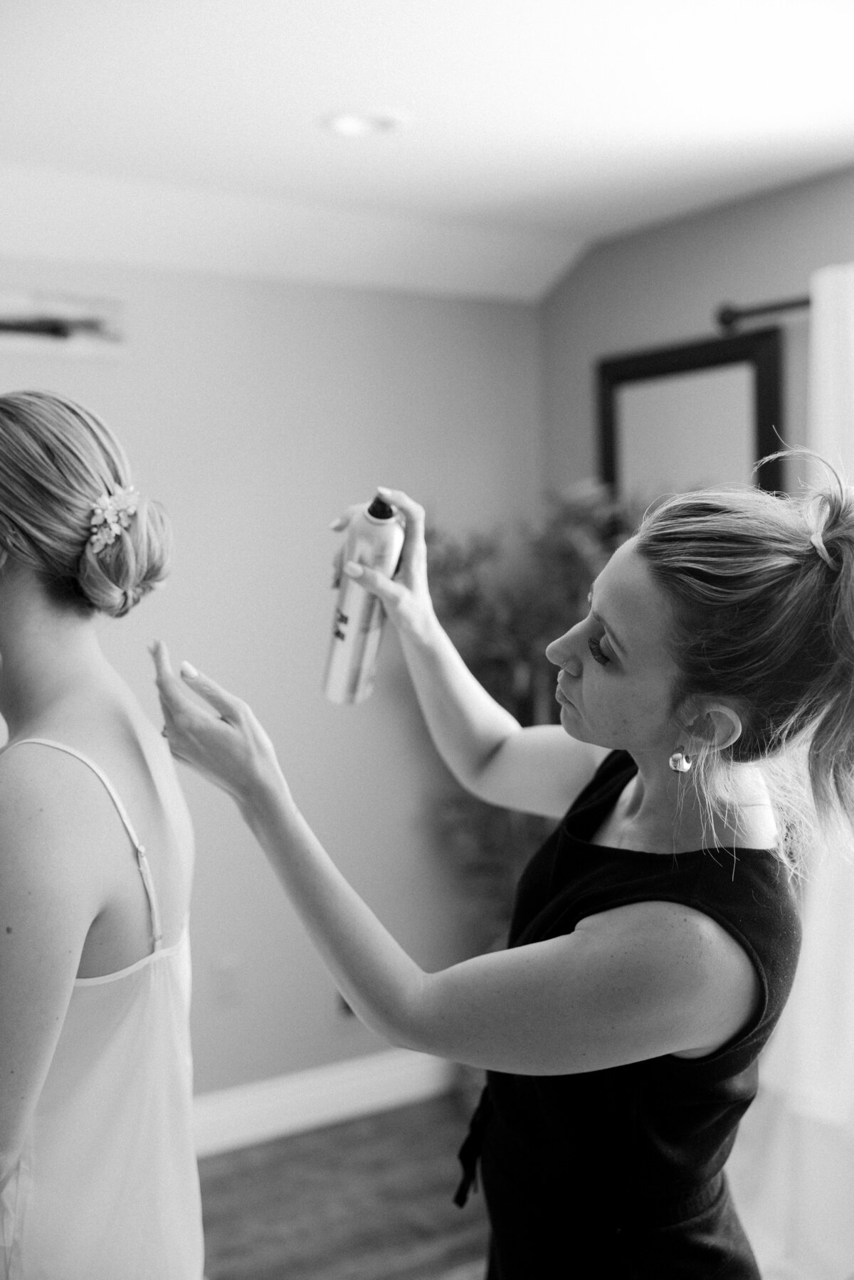 Black and white image of Amy from promise bridal artistry hair spraying the final touches of her bride's hair. 