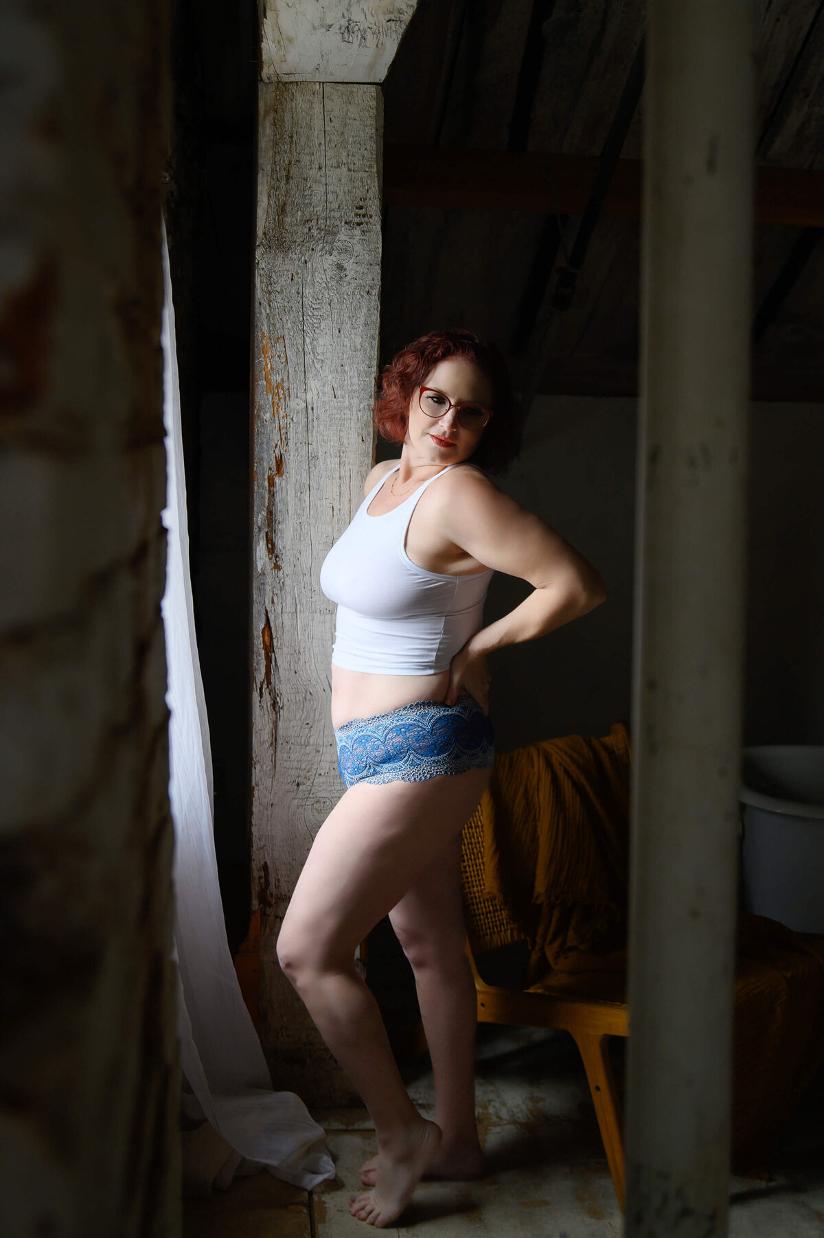Red haired woman in white crop tank and blue underwear with red glasses standing by a wood beam for her Hamilton boudoir photography session.