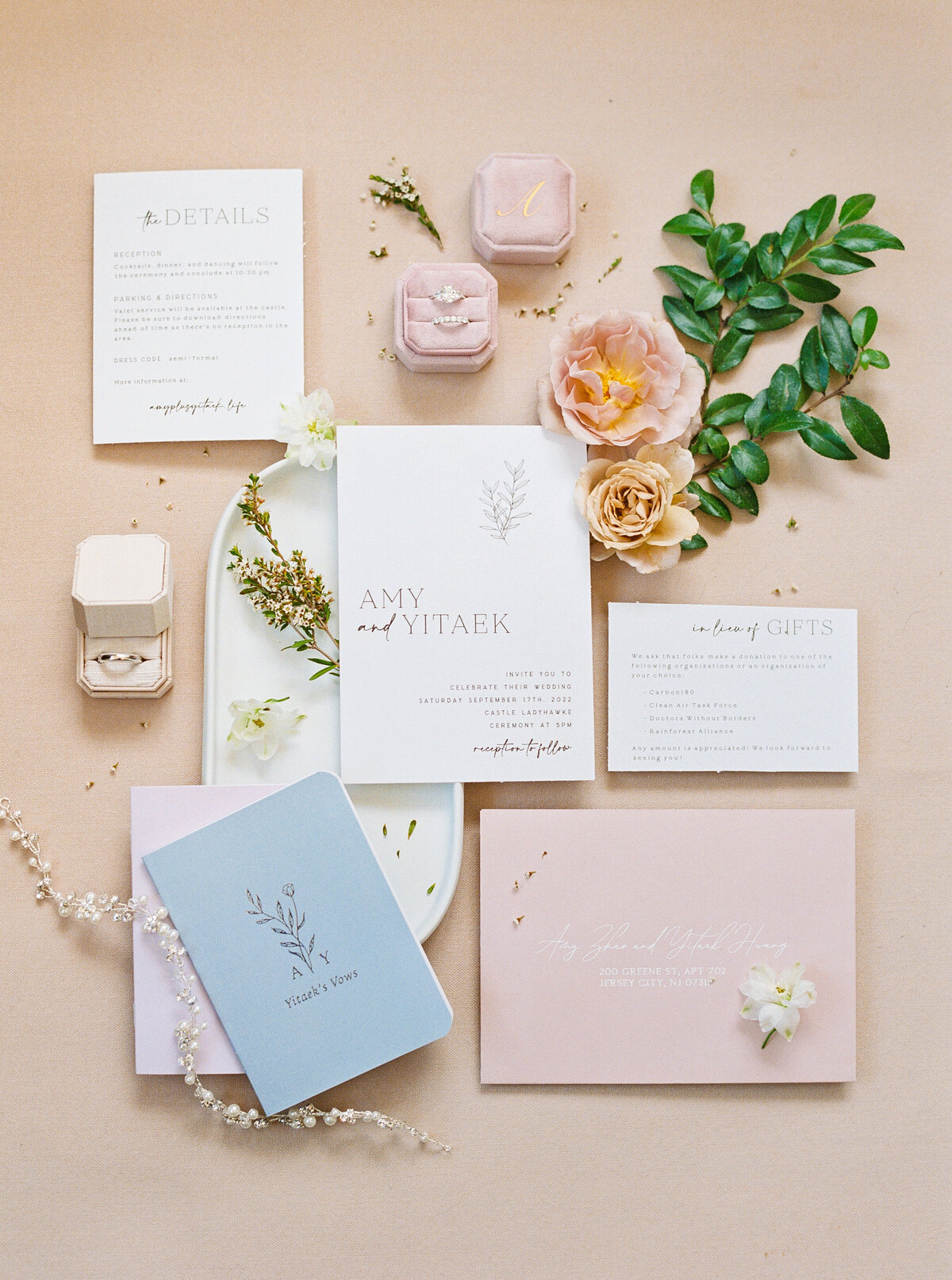 Flatlay of pastel wedding invitations, vow books, and ring boxes surrounded by florals at Castle Ladyhawke in Tuckaseegee, NC.