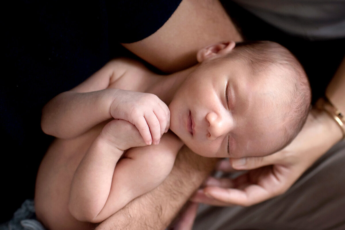 A newborn boy clasping his hands while sleeping in his father's arms at his newborn session in Dallas, TX.