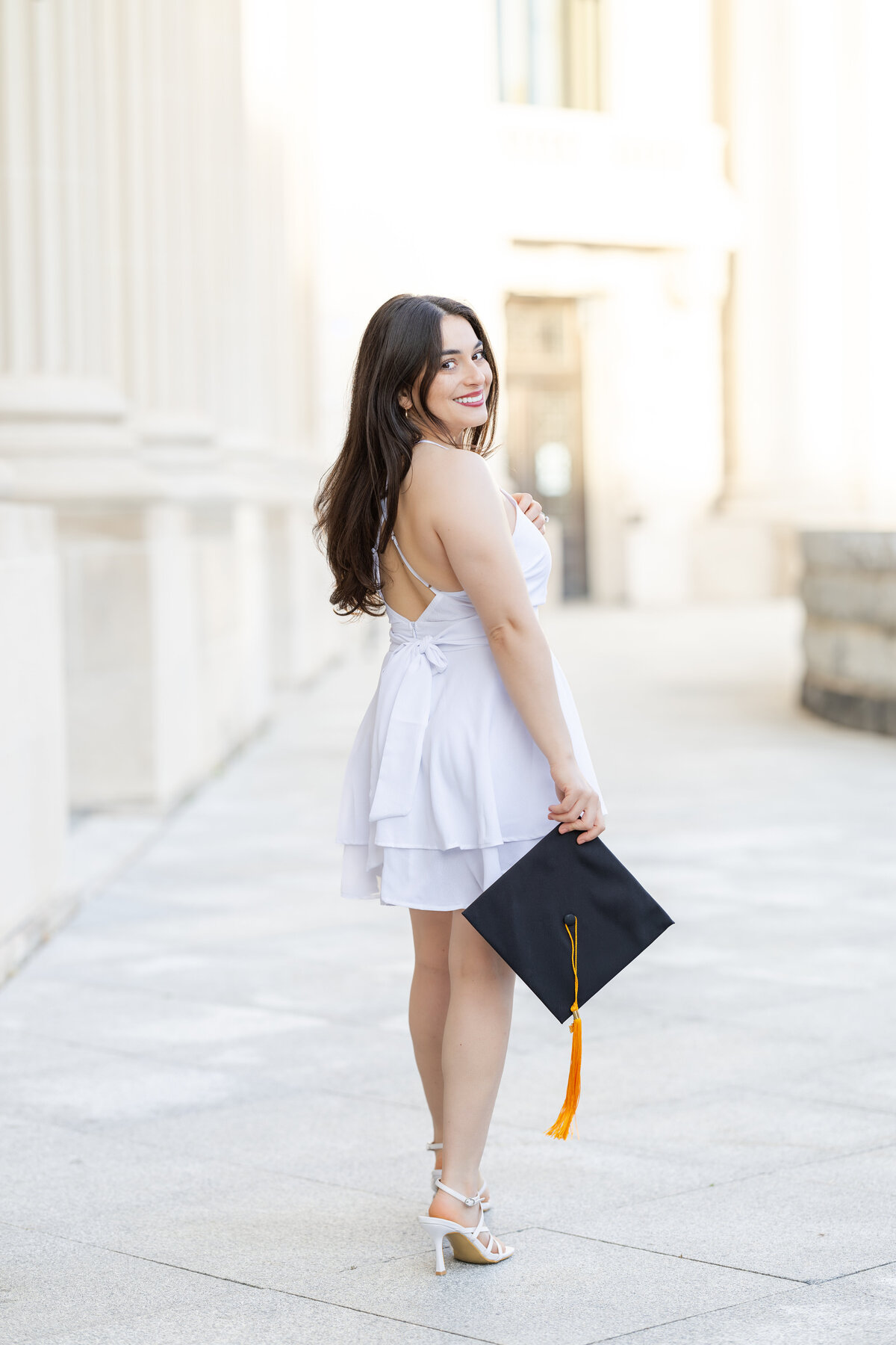 A young woman with long dark hair stands on a grand outdoor walkway lined with tall columns, wearing a white, tiered, backless dress and white heels.  She holds a black graduation cap with a gold tassel in her hand while smiling over her shoulder at the camera. 