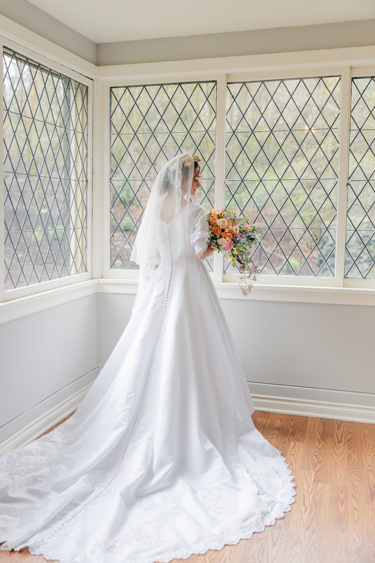 Bride softly looks down towards her spring colorful bouquet and her satin wedding gown trails the floor of the glover mansion