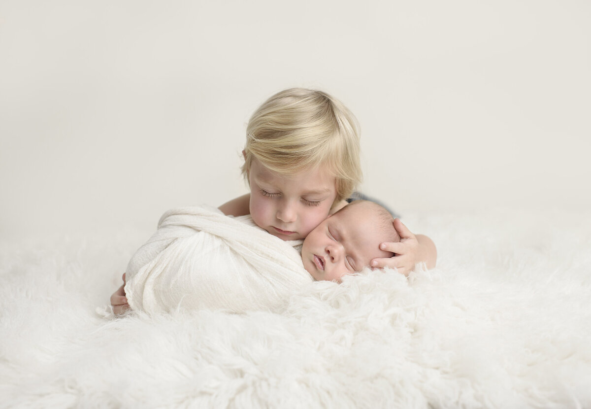 brother sitting with newborn baby sibling thats wrapped in white blanket and a white backdrop
