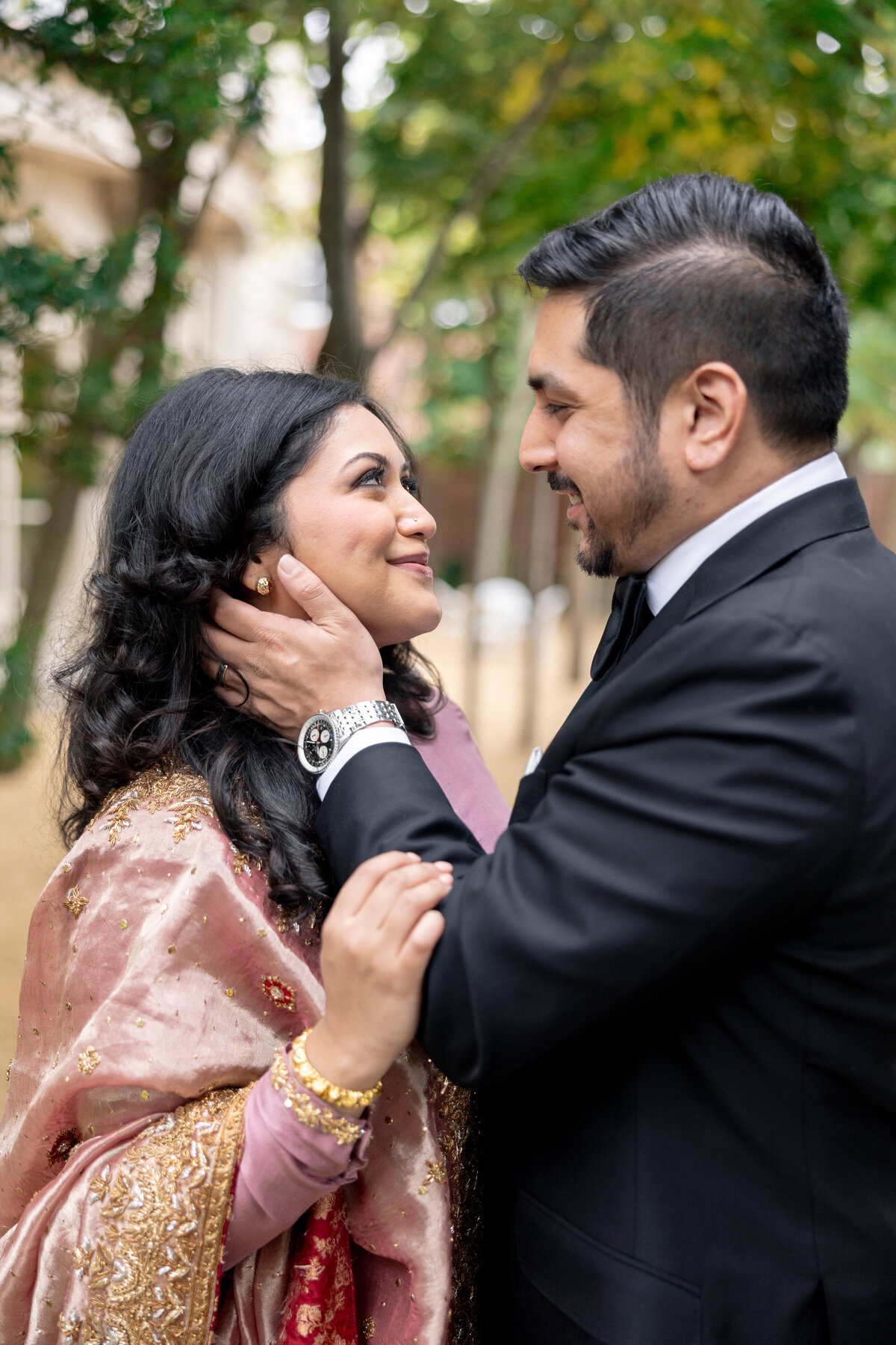 A couple stands outdoors, gazing into each others eyes. The woman has long dark hair and wears a vibrant red and gold outfit. The man, with short dark hair and a beard, is in a dark suit. They both smile affectionately.