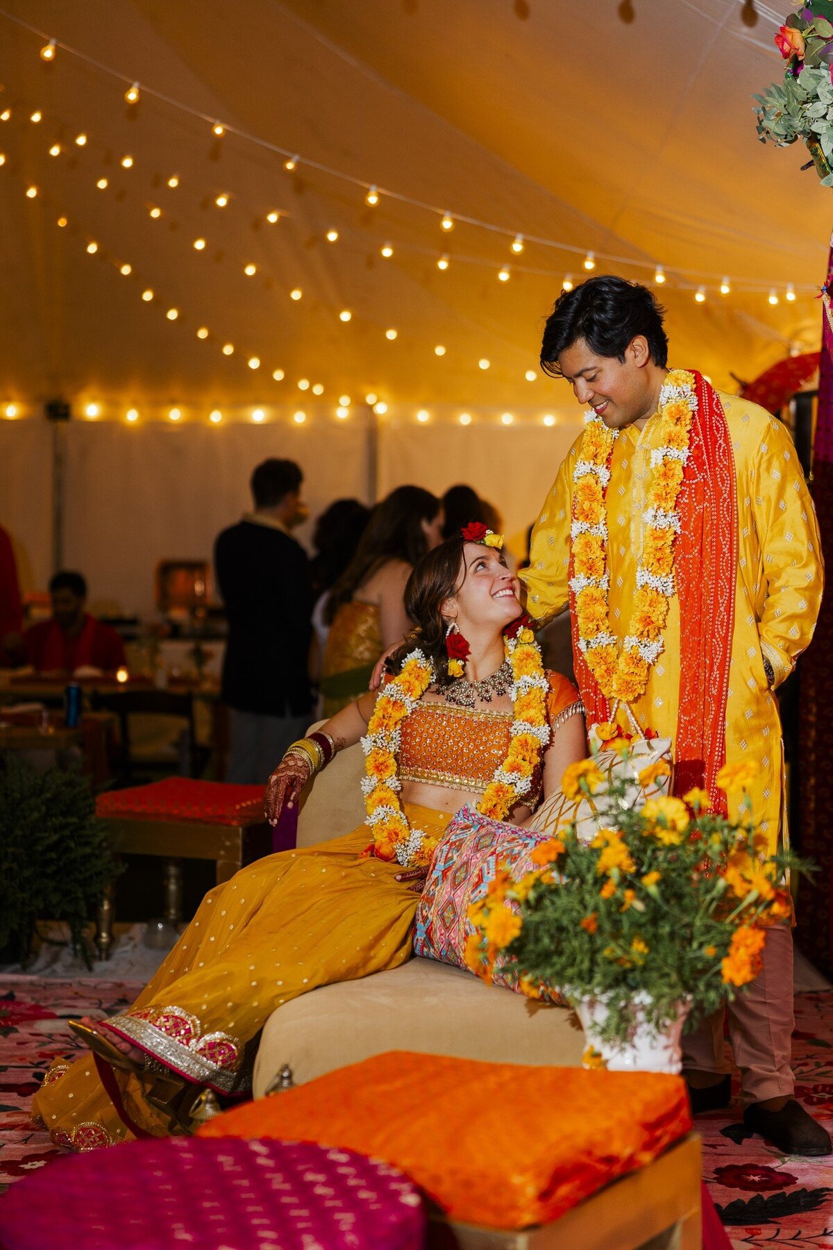 Couple in bright yellow outfits with marigold garlands seated together at a South Asian wedding welcome party