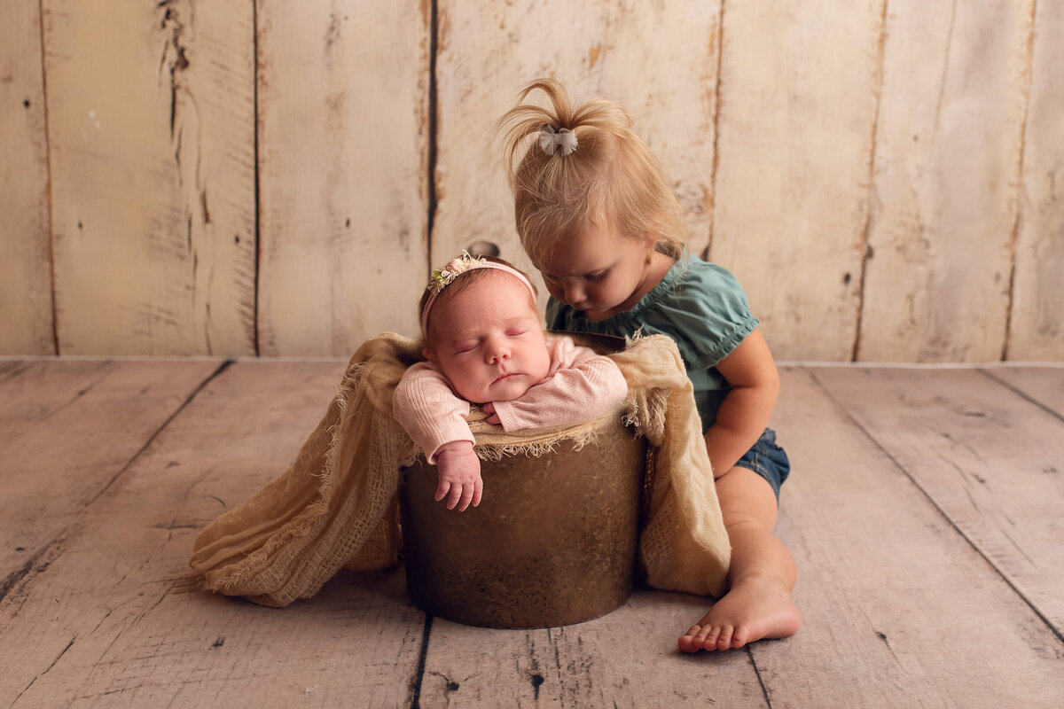 Neutral-colored portrait of newborn and toddler sisters in Waukesha photo studio. Baby is sleeping in a bucket and sister is crouched next to her watching intently.