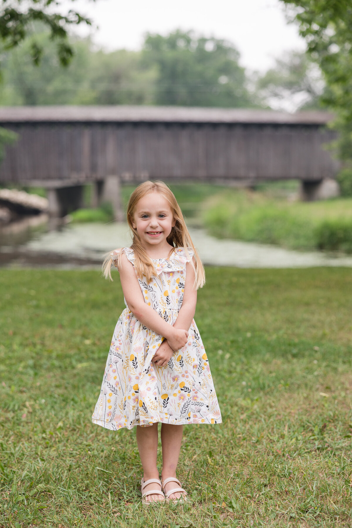 A little girl is standing in front of a covered bridge smiling at the camera