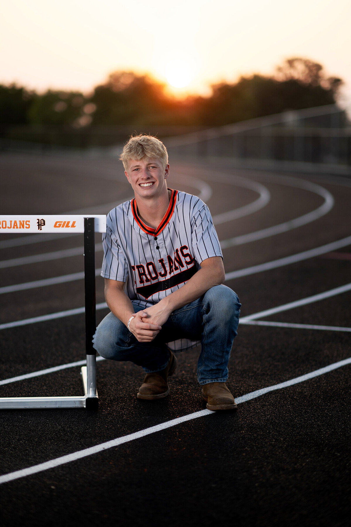 A Pella Senior Photography image of a high school senior squatting and smiling on a track with a hurdle
